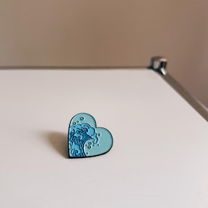 Könnte beinhalten: Herzförmige Emaille-Anstecknadel mit hellblauem Hintergrund und dunkelblauem Wellenmuster. Die Anstecknadel hat einen schwarzen Rand. Dekoratives Accessoire.