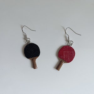 May include: A pair of earrings featuring miniature ping pong paddles. One paddle is black and the other is red.