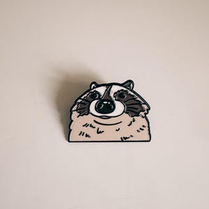 Könnte beinhalten: Ein Emaille-Pin in Form eines Waschbären mit schwarzer Umrandung. Der Pin hat ein weißes Gesicht mit schwarzen Augen und einer schwarzen Nase. Das Fell des Waschbären ist beige und braun. Der Pin befindet sich auf weißem Hintergrund.