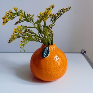 Può includere: Un vaso di ceramica a forma di arancia con un dettaglio di foglia verde, che contiene un mazzo di fiori gialli.