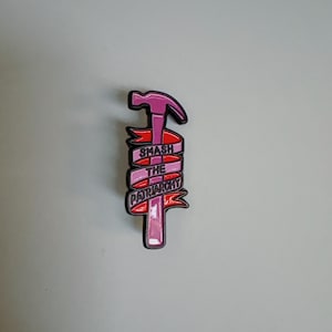 Könnte beinhalten: Ein pinkfarbener und roter Emaille-Pin in Form eines Hammers. Ein rotes Banner mit dem weißen Text "SMASH THE PATRIARCHY" umschließt den Hammer. Der Pin hat eine schwarze Umrandung und ist vor einem hellgrauen Hintergrund platziert.