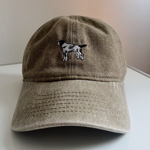 Könnte beinhalten: Eine braune Baseballkappe aus Canvas mit einem weißen gestickten Bild eines Hundes auf der Vorderseite.