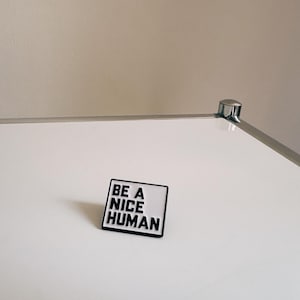May include: A rectangular pin with the text "BE A NICE HUMAN" in black letters on a white background, framed by a black border. The pin is set against a white surface.