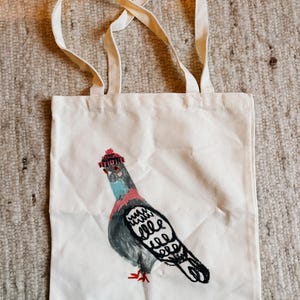May include: White tote bag with a hand-stitched design of a pigeon wearing a red knit cap. The pigeon is gray, black, and pink, with a blue neck. The bag has two cream-colored handles.