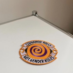 Könnte beinhalten: Gestickter Aufnäher mit orangefarbenem Hintergrund und einem lila Spiraldesign. Der Aufnäher trägt die Aufschrift "CINNAMON ROLLS" über der Spirale und "NOT GENDER ROLES" darunter. Der Aufnäher hat einen weißen Rand und liegt auf einer weißen Oberfläche.