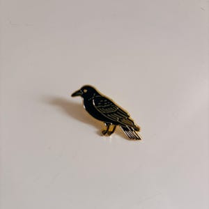 May include: A black raven pin with gold-colored trim. The bird is depicted in a side profile, with its beak and eye visible. The pin is likely made of metal and has a glossy finish. The raven is a popular symbol in gothic and alternative fashion.