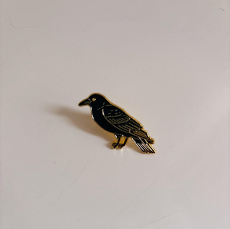 May include: A black raven pin with gold-colored trim. The bird is depicted in a side profile, with its beak and eye visible. The pin is likely made of metal and has a glossy finish. The raven is a popular symbol in gothic and alternative fashion.