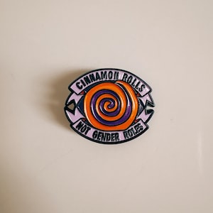 Könnte beinhalten: Emaille-Anstecknadel mit einem Zimtschnecken-Design. Die Anstecknadel zeigt eine orangefarbene und violette Wirbel im Zentrum, umgeben von den Worten "CINNAMON ROLLS" und "NOT GENDER ROLES" in schwarzem Text auf hellviolettem Hintergrund.