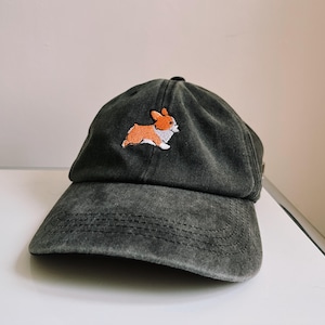 Könnte beinhalten: Eine dunkelgraue Baseballkappe mit einem kleinen gestickten Bild eines Corgi-Hundes in Orange und Weiß.