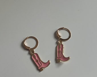 Pode incluir: Um par de brincos de argola dourados com pingentes de botas de cowboy rosa. As botas têm uma estrela dourada na frente.