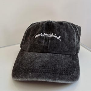 May include: A dark gray baseball cap with the word "overstimulated" embroidered in white script across the front. The cap has a curved brim and a distressed, washed appearance.