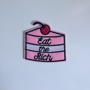 Könnte beinhalten: Rosa gestickter Bügel-Patch mit einem Kuchen-Design und dem Text "Eat the Rich".