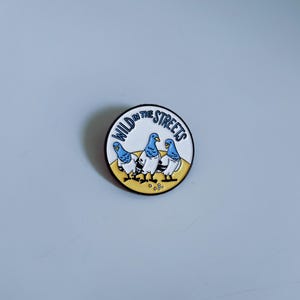 Könnte beinhalten: Eine weiße Emaille-Anstecknadel mit gelbem Hintergrund und drei blauen Tauben. Der Text "WILD IN THE STREETS" ist in einem schwarzen Kreis um die Tauben geschrieben.