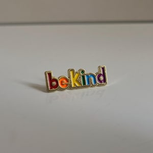 May include: A gold-colored pin with the words "be kind" in rainbow colors. The pin has a gold border and is set against a white background. The letters are in red, orange, yellow, green, blue, and purple.