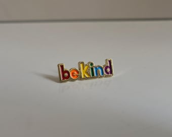 Könnte beinhalten: Eine goldfarbene Anstecknadel mit dem Schriftzug "be kind" in Regenbogenfarben. Die Anstecknadel hat einen goldenen Rand und ist vor einem weißen Hintergrund platziert. Die Buchstaben sind rot, orange, gelb, grün, blau und lila.