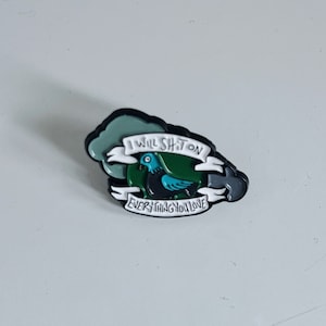 Op de afbeelding: Een zwart-witte emaille pin met een groene en blauwe vogel en de tekst "I will shit on everything you love."