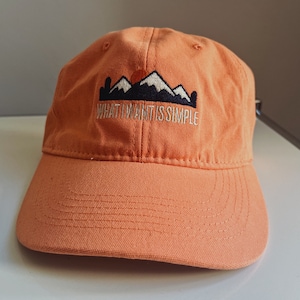 Könnte beinhalten: Eine orangefarbene Baseballkappe mit einer weißen gestickten Bergkette und dem Text "WHAT I WANT IS SIMPLE".