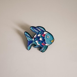 May include: A colorful fish-shaped decorative item. The fish features a blue body with a variety of scales in shades of blue, white, pink, and teal. It has a yellow mouth and a black outline.