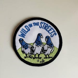 Op de afbeelding: Een witte geborduurde patch met een zwarte rand. De patch toont drie duiven met blauwe koppen en vleugels, en de tekst "WILD IN THE STREETS" in blauw.
