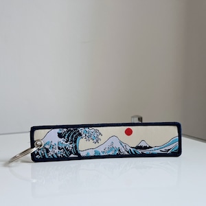 May include: A blue and white keychain featuring a depiction of The Great Wave off Kanagawa, a famous Japanese woodblock print by Hokusai. The keychain includes a red sun and a black border.
