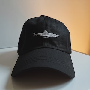 Könnte beinhalten: Eine schwarze Baseballkappe mit einem weißen gestickten Hai-Design.