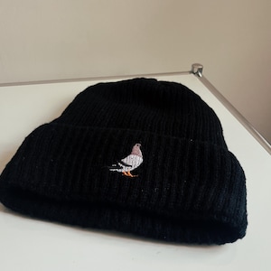 Könnte beinhalten: Schwarze Strickmütze mit einem weißen gestickten Taubenmotiv.