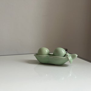 Puede incluir: Un juego de salero y pimentero de cerámica con forma de guisantes en una vaina. Los saleros son de color verde claro y tienen un acabado brillante.