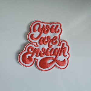 Könnte beinhalten: Ein weißer Patch mit roter Stickerei, auf dem "You are enough" steht.