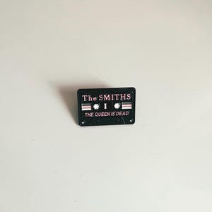 Puede incluir: Pin de esmalte negro con forma de casete con el texto "The Smiths" y "The Queen Is Dead" en letras rosas.