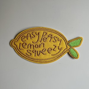 Könnte beinhalten: Gelber gestickter Bügelaufkleber in Form einer Zitrone mit dem Text "Easy Peasy Lemon Squeezy" in braunem Garn.