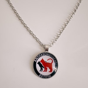 Op de afbeelding: Een zilveren ketting met een ronde hanger met een zwart en rood katten silhouet met de tekst "ANTIFASCHISTISCHE AKTION" in zwart en wit.