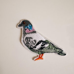 Könnte beinhalten: Gestickter Aufnäher eines Tauben. Der Vogel hat einen grauen Kopf, einen weißen Körper und grüne und schwarze Flügel. Der Aufnäher hat orangefarbene Füße und einen rosa und blauen Fleck am Hals.