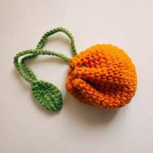 Puede incluir: Una bolsa naranja de ganchillo hecha a mano con detalles de tallo y hojas verdes. La bolsa es de color naranja vibrante, y el tallo y la hoja son de un verde brillante. La bolsa probablemente se usa para objetos pequeños o como pieza decorativa.