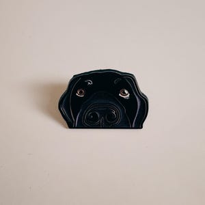 Könnte beinhalten: Ein schwarzes, hundeförmiges Dekorationsobjekt mit einer detaillierten Illustration des Gesichts eines Hundes. Der Hund hat braune Augen und eine detaillierte Nase. Der Artikel ist wahrscheinlich eine Anstecknadel, ein Magnet oder ein Aufkleber.