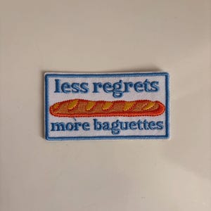 Könnte beinhalten: Ein rechteckiger bestickter Aufnäher mit weißem Hintergrund und blauem Rand. Der Text "less regrets" steht in Blau über einer goldbraunen Baguette-Illustration mit orangefarbenem Umriss, gefolgt von dem Text "more baguettes" in Blau.