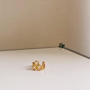 Könnte beinhalten: Ein goldfarbener Ring mit einer Reihe von Smiley-Gesicht-Ausschnitten. Der Ring besteht aus glänzendem Metall und liegt auf einer weißen Oberfläche.