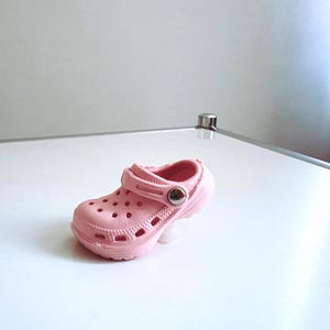Könnte beinhalten: Ein einzelner rosa Clog-Schuh mit Riemen und mehreren Belüftungslöchern. Der Schuh besteht aus einem gummiartigen Material und hat einen kleinen silbernen Knopf am Riemen. Der Schuh ist für Kinder konzipiert.