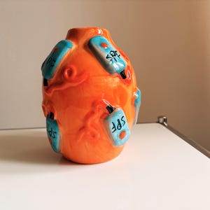 Könnte beinhalten: Eine leuchtend orangefarbene Keramikvase mit erhabenen, dreidimensionalen hellblauen Flaschenformen auf der Oberfläche. Jede blaue Flasche ist mit dem schwarzen Text SPF beschriftet und hat ein kleines schwarzes Verschlussdetail.