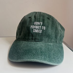 May include: A green, distressed, cotton baseball cap with white embroidered text that reads "Don't forget to smile".