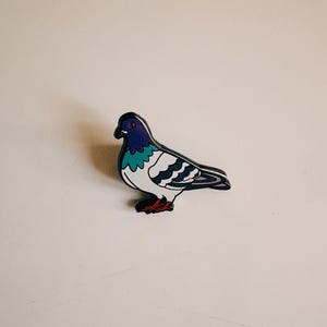 Könnte beinhalten: Eine farbenfrohe, taubenförmige Anstecknadel mit schwarzer Umrandung. Die Anstecknadel hat einen lila Kopf, eine türkisfarbene Brust, einen weißen Körper und schwarze und graue Flügel. Die Füße der Taube sind rot.