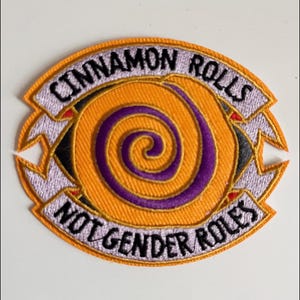 Puede incluir: Parche bordado con el texto "CINNAMON ROLLS" arriba y "NOT GENDER ROLES" debajo de un diseño en espiral. El parche presenta un fondo naranja con una espiral morada y una pancarta blanca con texto negro.