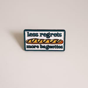 May include: A rectangular pin with a white background and a teal border. The pin features the text "less regrets" above an illustration of a baguette, and the text "more baguettes" below.