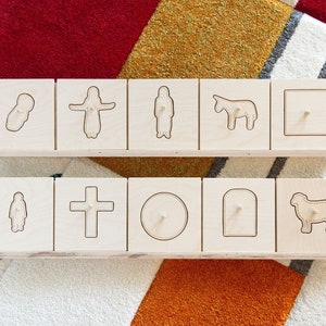 May include: A set of wooden blocks with various shapes and symbols, including a cross, a circle, a rectangle, a person, a sheep, a donkey, and a figure in a robe. The blocks are arranged in two rows of six, with the top row featuring the symbols and the bottom row featuring the shapes.