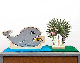 May include: A gray wooden whale with a yellow fish in its mouth. The whale is on a blue surface and there is a wooden flower vase with pink flowers and a green palm leaf on a green surface.