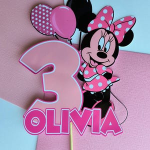 Op de afbeelding: Roze en zwarte Minnie Mouse taarttopper met een roze nummer 3, ballonnen en de naam Olivia.