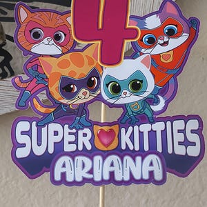 May include: A vibrant cake topper with the words "SUPER KITTIES ARIANA" and the number "4". It showcases cartoon cat characters dressed as superheroes. The topper is attached to a wooden stick.