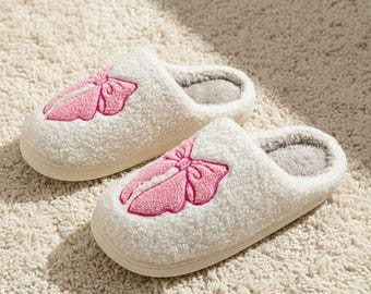 Può includere: Un paio di pantofole bianche e soffici con un design a fiocco rosa sulla parte superiore. Le pantofole hanno un interno morbido e grigio e una suola color crema. Progettate per l'uso in casa, sono posizionate su un tappeto color crema.