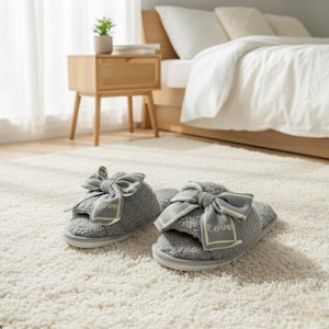 Op de afbeelding: Een paar grijze slippers met open teen, een zachte textuur en een decoratieve strik. De strik heeft een lichtgrijs lint met het woord "Love" erop gedrukt. De slippers liggen op een wit tapijt.