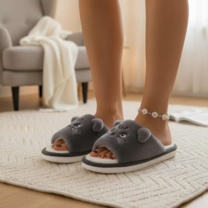 May include: Gray plush slippers with a cartoon cat face design. The slippers have a white sole with a gray stripe and are open-toed. The image shows the slippers being worn indoors on a light-colored rug.