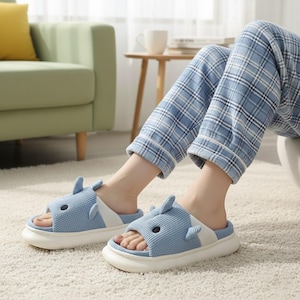 May include: Light blue shark-themed slippers with white accents and a thick white sole. The open-toe slippers have a textured waffle-weave design and small fins. The slippers are worn with blue plaid pajama pants.